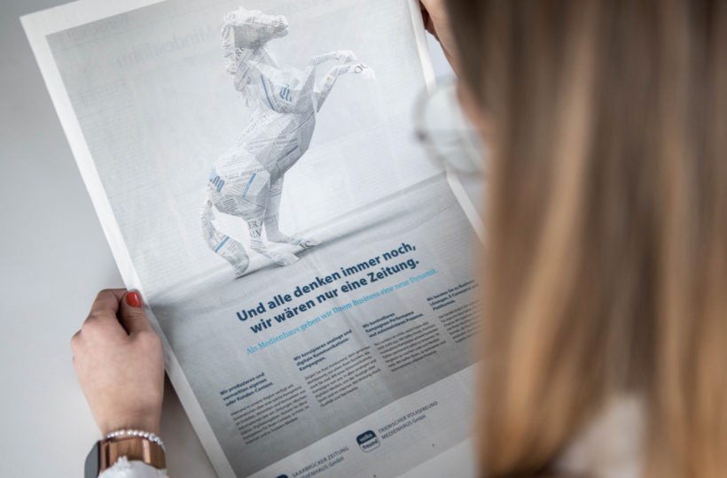 Eine Person mit langen Haaren und Brille hält eine Zeitung in der Hand, auf deren Seite eine Pferdeskulptur aus Zeitungspapier abgebildet ist, darunter ein Text in deutscher Sprache mit einer fettgedruckten Schlagzeile.