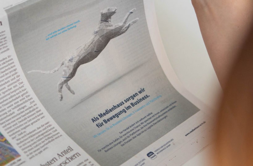 Eine Person hält eine Zeitung in der Hand, die eine Anzeige mit einer springenden Hundefigur aus Zeitungsseiten und deutschem Text über Mediendienste, Wirtschaft und Bewegung zeigt.