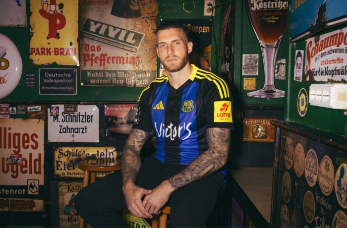 Ein Mann mit Tattoos sitzt während eines Fotoshootings auf einem Holzhocker in einer schummrigen, mit deutschen Schildern und Erinnerungsstücken dekorierten Bar im Vintage-Stil. Er trägt ein schwarz-blau gestreiftes Fußballtrikot mit gelben Streifen und Sponsorenlogos.