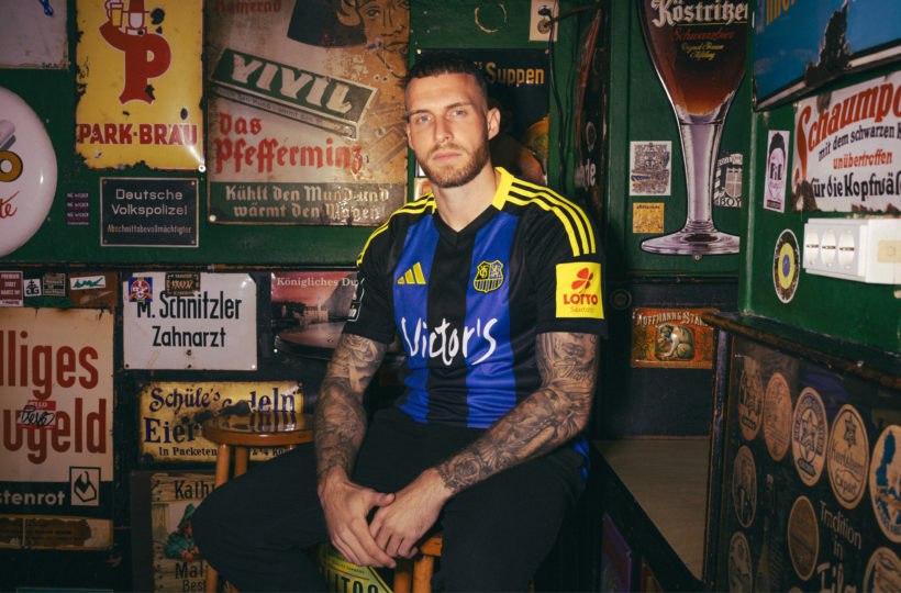 Ein Mann mit Tattoos sitzt während eines Fotoshootings auf einem Holzhocker in einer schummrigen, mit deutschen Schildern und Erinnerungsstücken dekorierten Bar im Vintage-Stil. Er trägt ein schwarz-blau gestreiftes Fußballtrikot mit gelben Streifen und Sponsorenlogos.