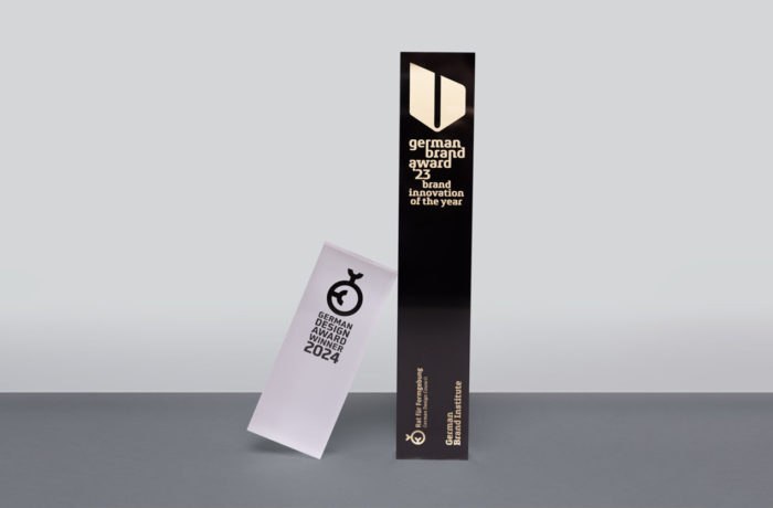 Zwei Designpreise stehen auf einer grauen Fläche: eine hohe, schwarze Trophäe des German Brand Award 2023 und eine kürzere, schräge weiße Plakette des German Design Award Special 2024. Beide zeichnen sich durch minimalistisches Design und einen fettgedruckten Text aus, der herausragende Markenleistungen würdigt.