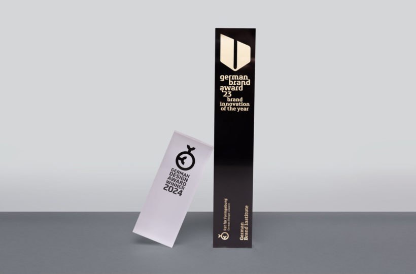 Zwei Designpreise stehen auf einer grauen Fläche: eine hohe, schwarze Trophäe des German Brand Award 2023 und eine kürzere, schräge weiße Plakette des German Design Award Special 2024. Beide zeichnen sich durch minimalistisches Design und einen fettgedruckten Text aus, der herausragende Markenleistungen würdigt.