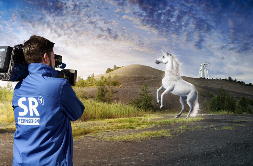 Ein Kameramann in einer blauen Jacke filmt ein sich aufbäumendes Einhorn auf einem grasbewachsenen Hügel unter einem teilweise bewölkten Himmel, mit einem weißen Industriegebäude im Hintergrund.