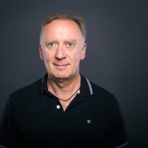 Ein Mann mittleren Alters mit kurzen hellbraunen Haaren und schwarzem Kragenhemd steht vor einem schlichten dunklen Hintergrund und blickt mit neutralem Blick in die Kamera.