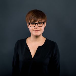 Eine Person mit kurzen braunen Haaren und schwarzer Brille, die ein schwarzes Oberteil trägt, steht vor einem schlichten dunklen Hintergrund und schaut mit neutralem Blick direkt in die Kamera.