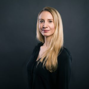 Eine Frau mit langen blonden Haaren, die eine schwarze Bluse trägt, steht vor einem dunklen, schlichten Hintergrund und lächelt sanft in die Kamera.