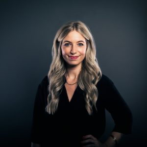 Eine Frau mit langem, gewelltem blondem Haar und schwarzem Outfit steht vor einem dunkelgrauen Hintergrund und lächelt selbstbewusst mit einer Hand in der Hüfte.