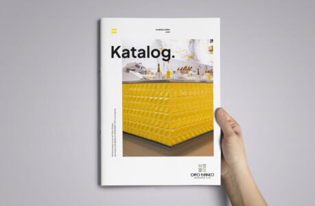 Eine Hand hält einen Katalog mit dem Titel "Katalog" in fetter schwarzer Schrift, der auf dem Umschlag einen modernen gelben Zähler mit geometrischem Muster zeigt. Das OroBianco-Logo befindet sich unten rechts.