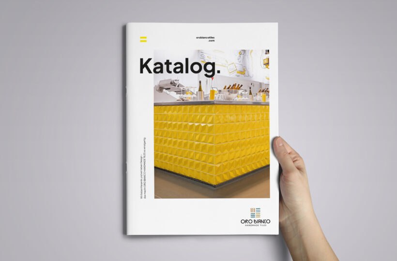 Eine Hand hält einen Katalog mit dem Titel "Katalog" in fetter schwarzer Schrift, der auf dem Umschlag einen modernen gelben Zähler mit geometrischem Muster zeigt. Das OroBianco-Logo befindet sich unten rechts.