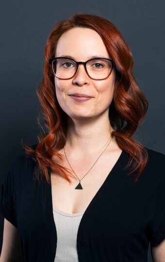 Franziska Himpel, eine Frau mit langen roten Haaren und Brille, die ein schwarzes Top über einem hellen Hemd und eine Dreieckskette trägt, steht vor einem schlichten dunklen Hintergrund und lächelt leicht.