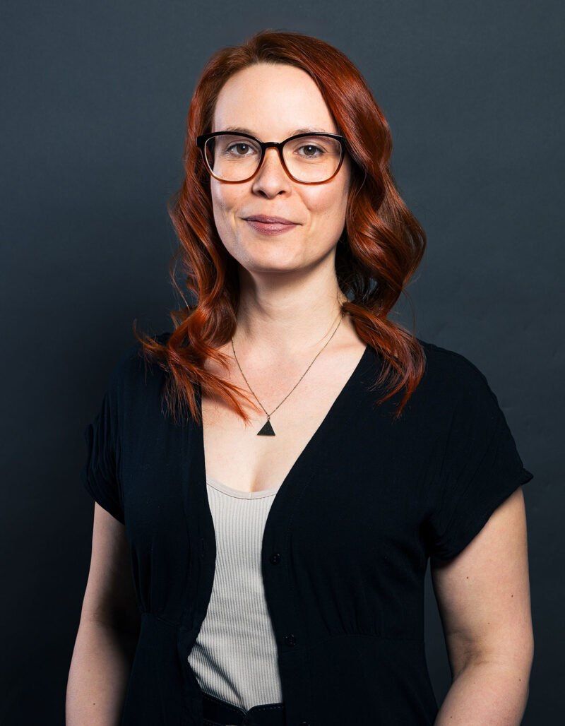 Franziska Himpel, eine Frau mit langen roten Haaren und Brille, die ein schwarzes Top über einem hellen Hemd und eine Dreieckskette trägt, steht vor einem schlichten dunklen Hintergrund und lächelt leicht.