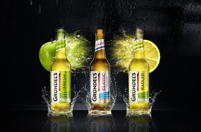 Drei CGI-Getränkeflaschen - Grundel's Fresh, Classic und Radler - stehen aufrecht auf einer nassen Oberfläche, um die Wasser spritzt. Fresh wird mit einem grünen Apfel und Radler mit einer Limettenscheibe kombiniert, alles vor einem dramatischen dunklen Hintergrund.