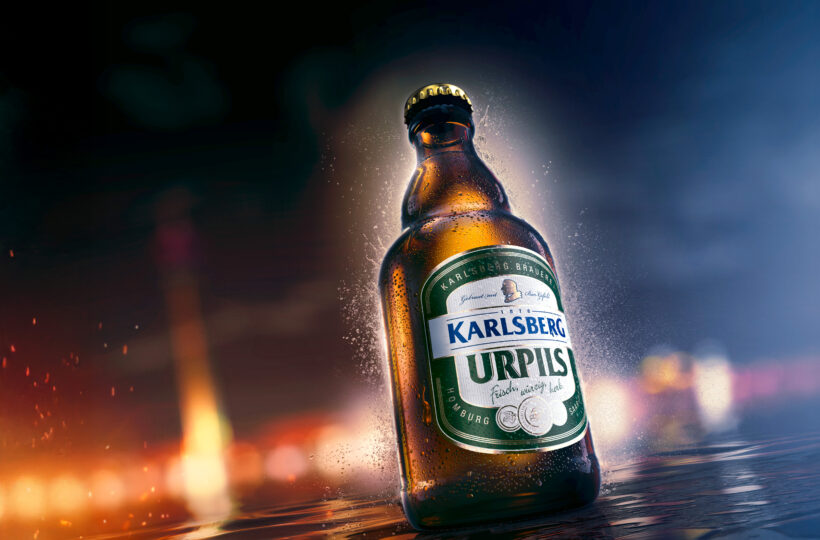 Eine kalte, braune Glasflasche Karlsberg Urpils Bier steht auf einer nassen, spiegelnden Oberfläche in dieser CGI-Getränkeszene, umgeben von Tropfen und vor einem unscharfen, farbenfrohen Hintergrund, der eine lebhafte Atmosphäre suggeriert.