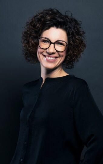 Eine sympathische und selbstbewusste Frau mit kurzen lockigen Haaren, lächelt, während sie vor einem dunklen, schlichten Hintergrund steht.