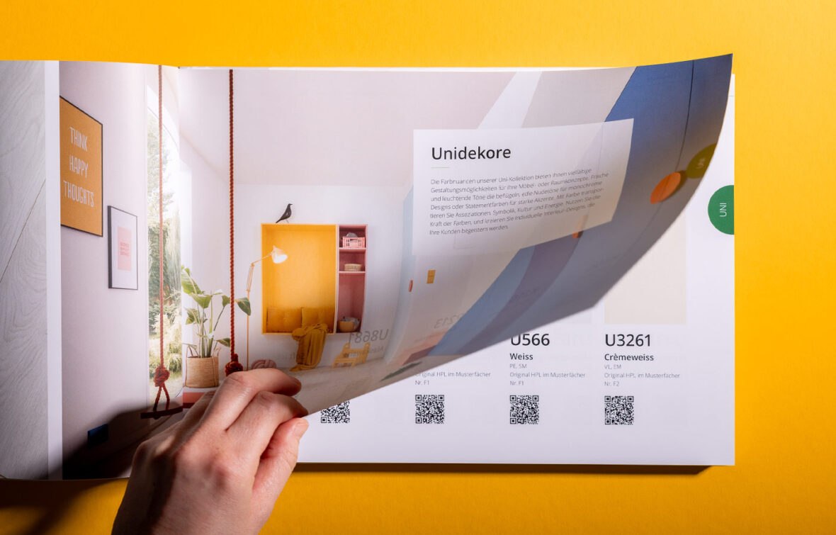 Eine Hand blättert auf einer leuchtend gelben Fläche durch einen Farbkatalog, der redaktionelle Gestaltungsdetails wie Raumdekor-Bilder, Farbmuster, QR-Codes und deutsche Texte zeigt.