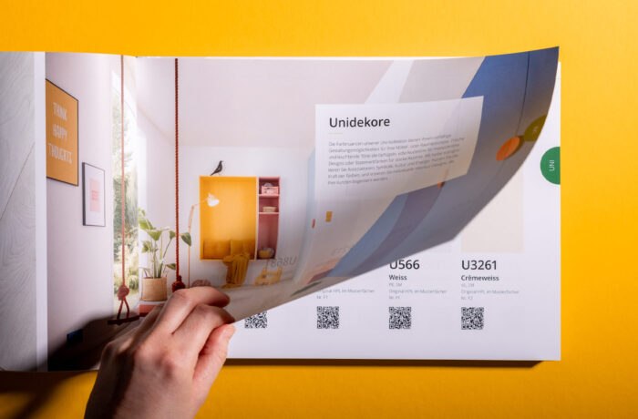 Eine Hand blättert auf einer leuchtend gelben Fläche durch einen Farbkatalog, der redaktionelle Gestaltungsdetails wie Raumdekor-Bilder, Farbmuster, QR-Codes und deutsche Texte zeigt.