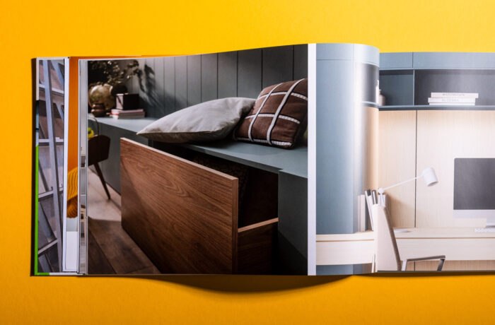 Ein aufgeschlagenes Magazin auf einer gelben Fläche zeigt moderne Einrichtungsfotos, eine Holzschublade mit Kissen und einen stilvollen Arbeitsbereich mit Regalen und einer Schreibtischlampe. Ein aufgeschlagenes Magazin auf einer gelben Fläche zeigt moderne Einrichtungsfotos, eine Holzschublade mit Kissen und einen stilvollen Arbeitsbereich mit Regalen und einer Schreibtischlampe.