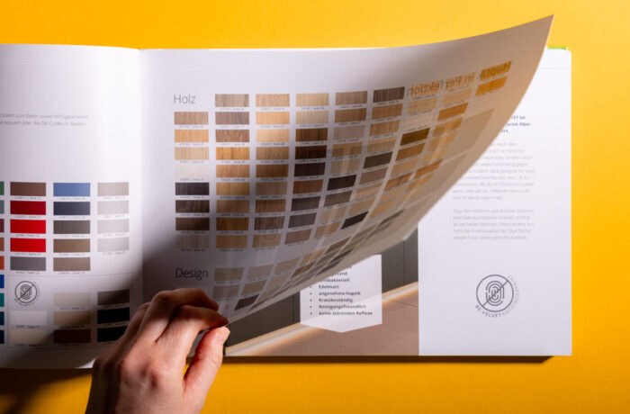 Eine Hand hebt eine transparente Seite in einer Musterbroschüre an, die das Editorial Design anhand verschiedener Holz- und Farbmuster mit beschreibendem Text in deutscher Sprache vor einem leuchtend gelben Hintergrund zeigt. Eine Hand hebt eine transparente Seite in einer Musterbroschüre an, die das Editorial Design anhand verschiedener Holz- und Farbmuster mit beschreibendem Text in deutscher Sprache vor einem leuchtend gelben Hintergrund zeigt.
