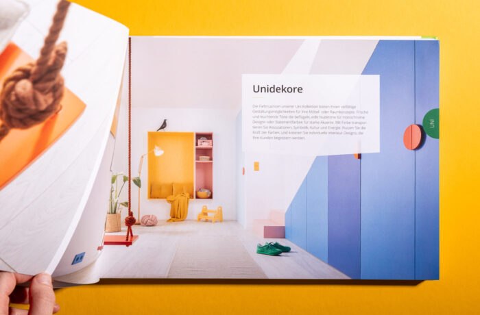 Ein aufgeschlagenes Buch auf einer gelben Fläche zeigt ein modernes, farbenfrohes Interieur mit rosa, orange, blau und weißem Dekor. Der deutsche Text steht in einem weißen Kasten am oberen Rand. Die Hand einer Person blättert eine Seite auf der linken Seite des Buches um. Ein aufgeschlagenes Buch auf einer gelben Fläche zeigt ein modernes, farbenfrohes Interieur mit rosa, orange, blau und weißem Dekor. Der deutsche Text steht in einem weißen Kasten am oberen Rand. Die Hand einer Person blättert eine Seite auf der linken Seite des Buches um.