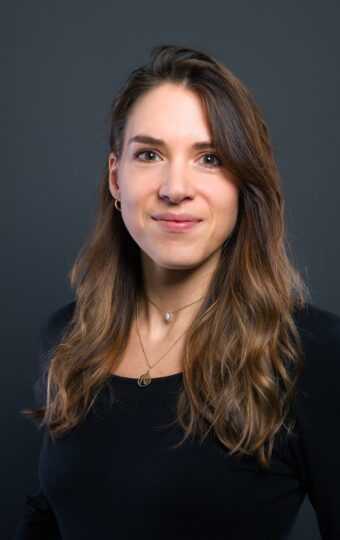 Nina Kronenberger, eine Frau mit langen braunen Haaren, die ein schwarzes langärmeliges Oberteil und eine Halskette trägt, steht vor einem schlichten dunklen Hintergrund und lächelt leicht in die Kamera.