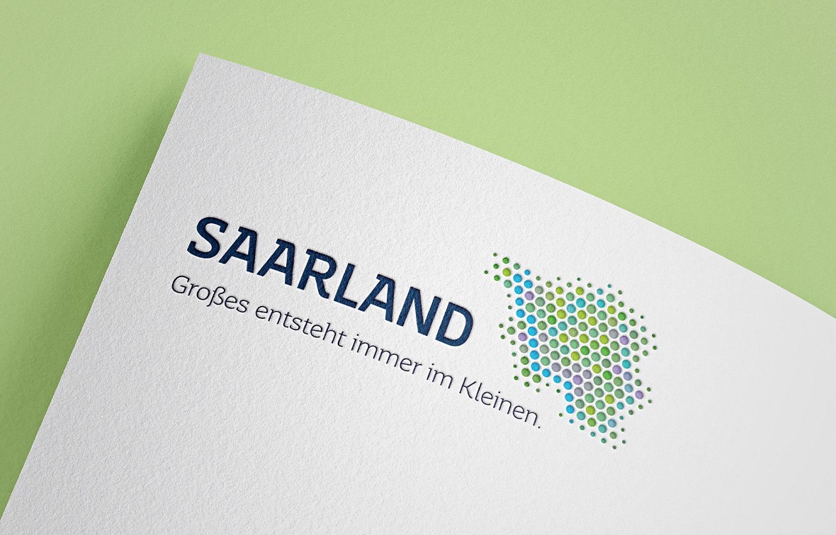 Ein von der HDW Werbeagentur Saarland entworfenes Logo auf weißem Papier zeigt das Wort SAARLAND, den Satz Großes entsteht immer im Kleinen und einen gepunkteten Kartenumriss in Grün, Blau und Lila auf hellgrünem Hintergrund.