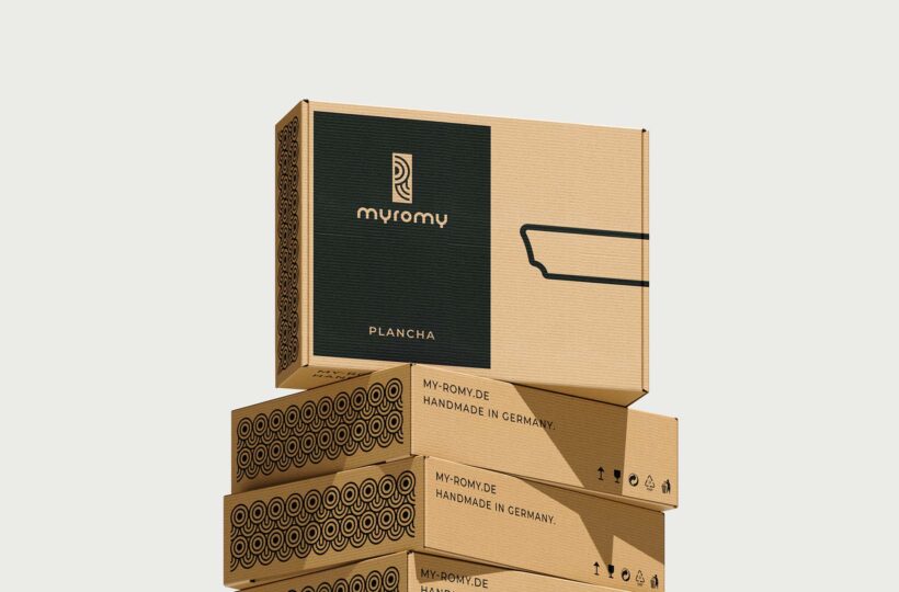 Ein Stapel von vier braunen Pappschachteln, wobei die oberste Schachtel mit dem myromy-Logo, dem Wort Plancha und einer einfachen Strichzeichnung versehen ist. Die schwarzen Muster und der Text "Handmade in Germany" unterstreichen Qualität und Barrierefreiheit.