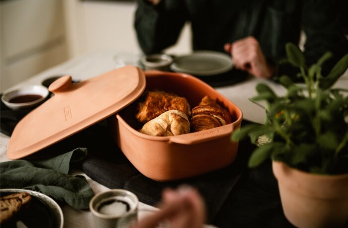 Eine Terrakottaschale mit Croissants und Gebäck steht auf einem Esstisch, umgeben von kleinen Schalen, Tellern und einer Topfpflanze - perfekt für eine Event-Kampagne zum Tag der deutschen Einheit, mit zwei Personen, die teilweise im Hintergrund zu sehen sind.