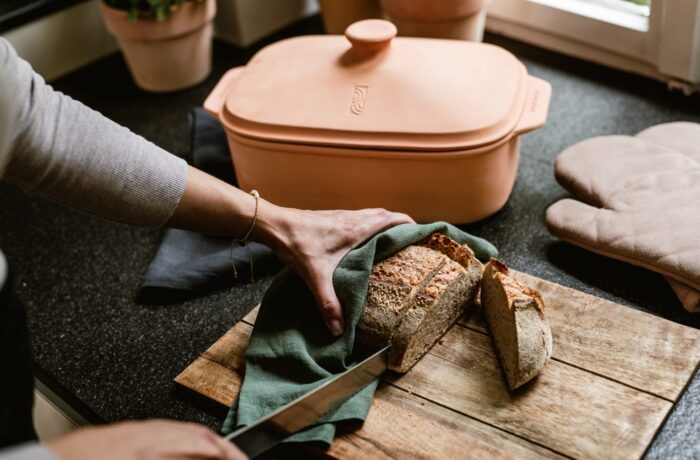 Während einer Event-Kampagne zum Tag der deutschen Einheit schneidet eine Person einen Laib Brot auf einem hölzernen Schneidebrett, das sie mit einem grünen Tuch abdeckt. Eine Terrakotta-Backform und Ofenhandschuhe liegen auf der schwarzen Arbeitsplatte daneben.